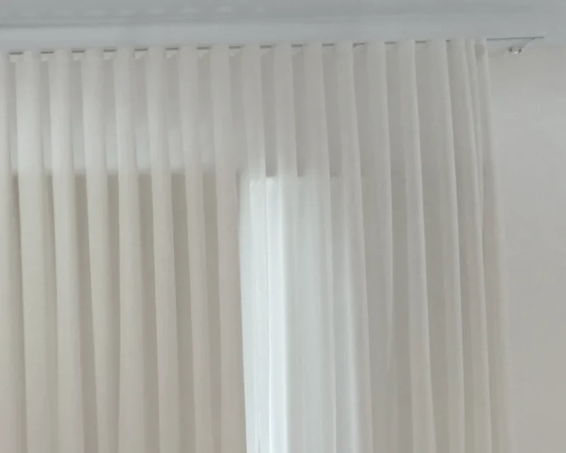 White sheer S-fold curtain hanging in even waves from a white ceiling track, with window light behind