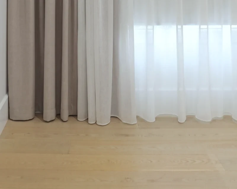 Double curtains at the floor: a white sheer in front of a taupe blockout, both hems touching a timber floor