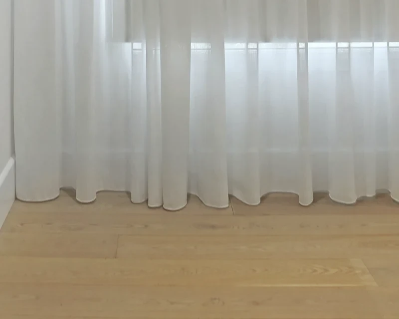 White sheer curtain hem just touching a timber floor, with daylight coming through the fabric