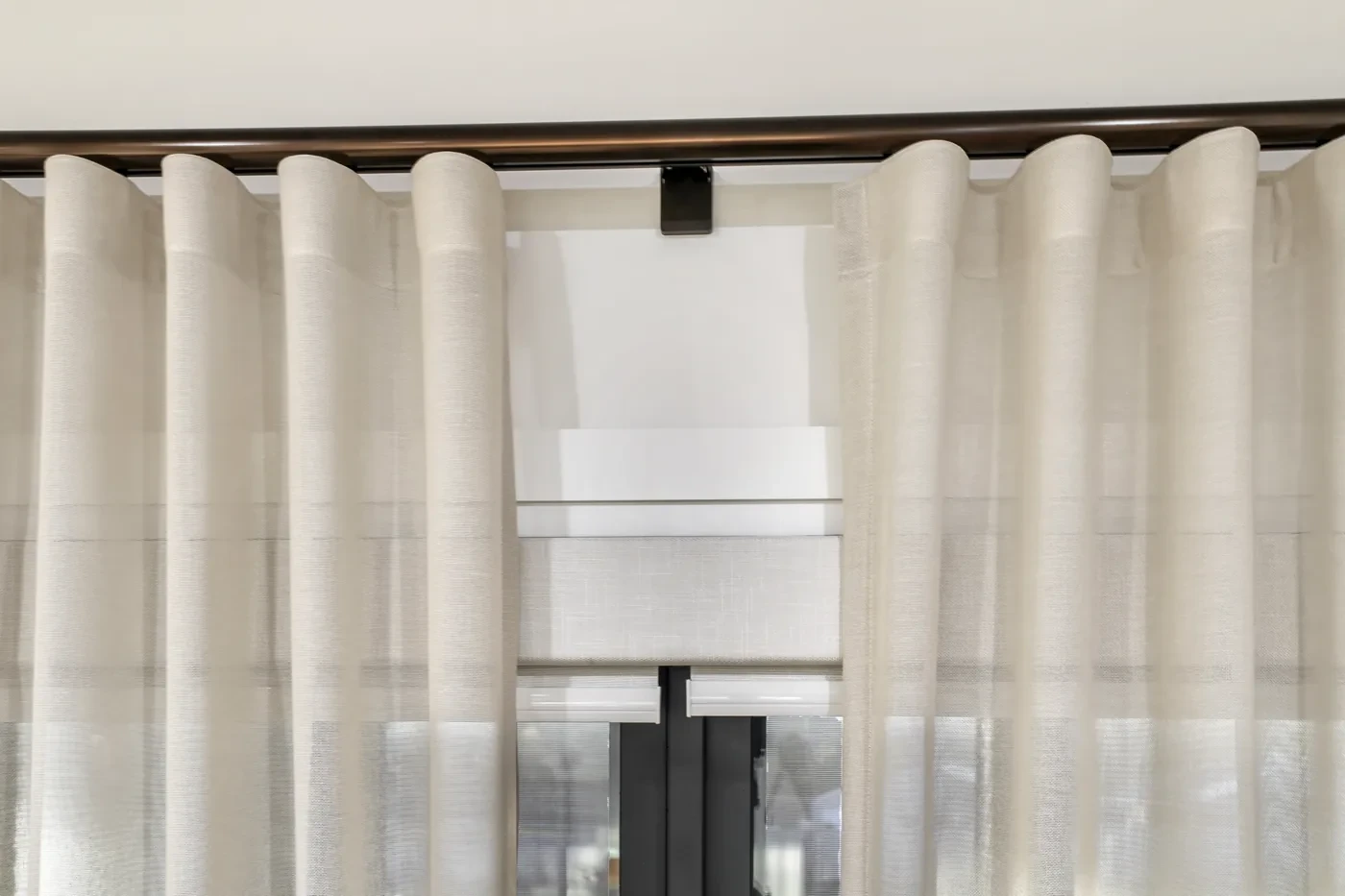 Close-up of a white blind layered behind S-fold sheer curtains on a dark rod Close-up of a white blind layered behind S-fold sheer curtains on a dark rod