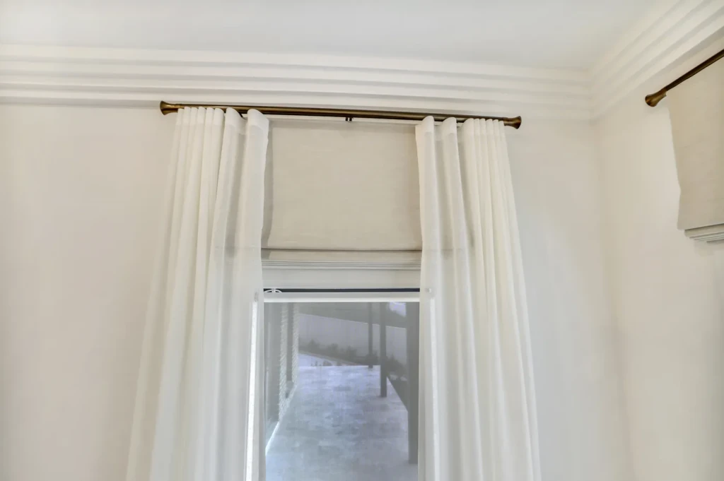 Light roman blind, part-raised so its folds show, layered under sheer curtains on a tall window