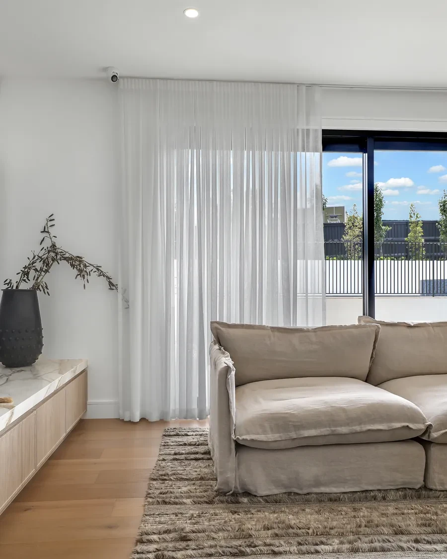 A white S-fold sheer curtain on a ceiling track drawn over the fixed glass beside a sliding door, behind a linen sofa