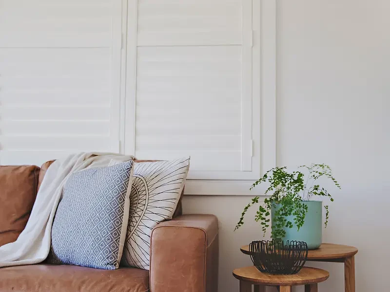 Closed white plantation shutters behind a tan leather sofa in a lounge room