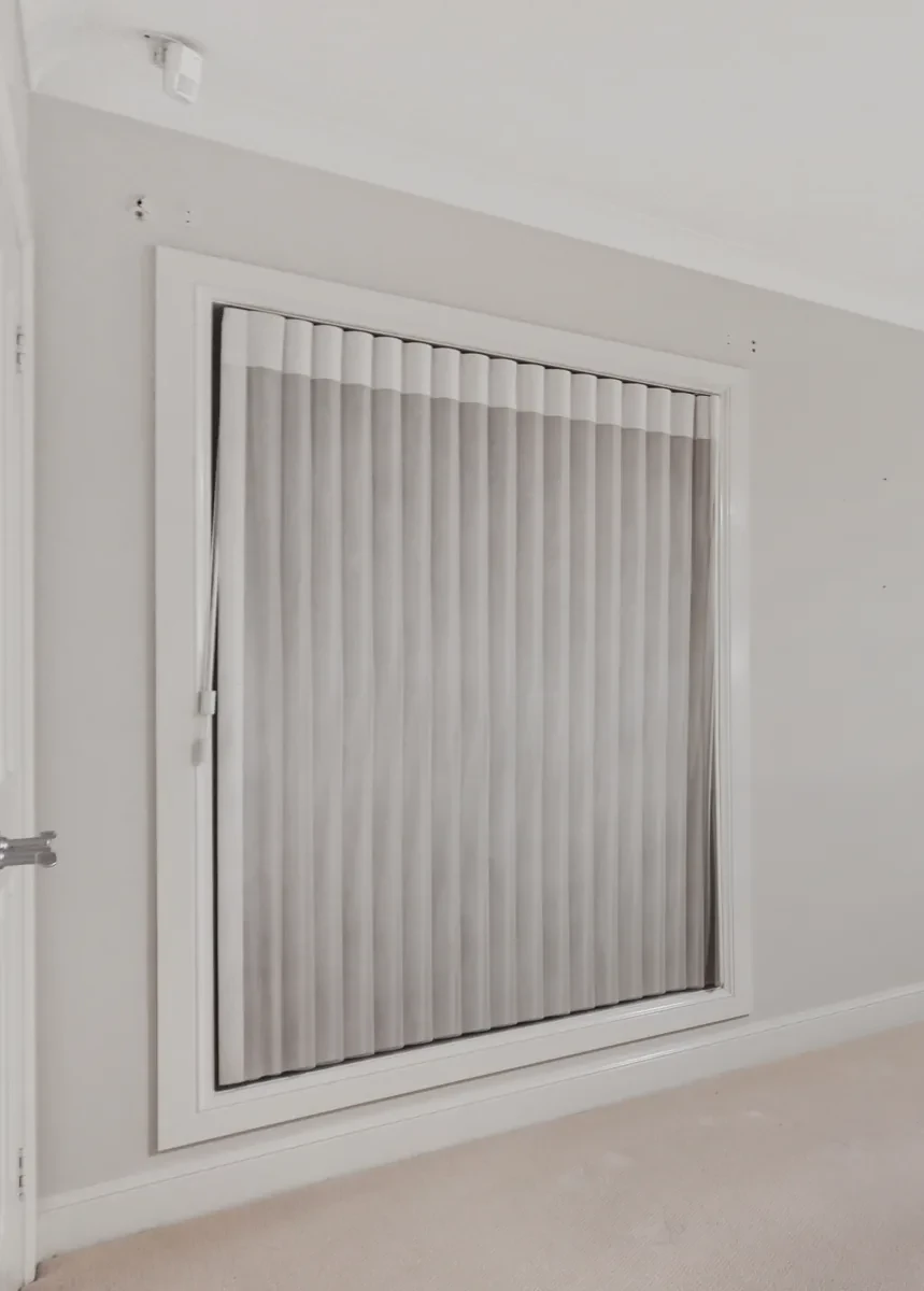 A grey fabric vertical blind fitted inside a white-framed bedroom window, vanes closed