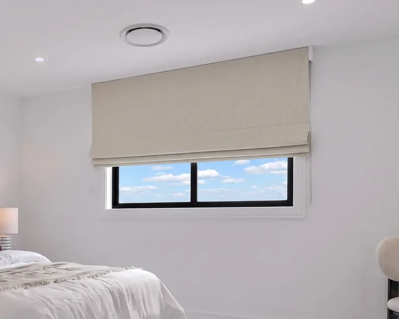 Oatmeal linen-look roman blind fixed to the wall above a bedroom window, covering the frame, part raised