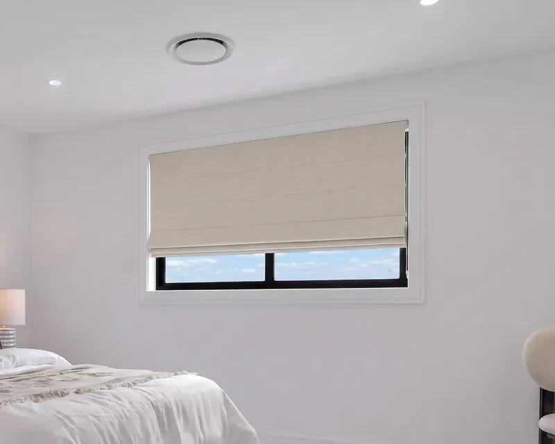 Oatmeal linen-look roman blind fitted inside a bedroom window recess, part raised with folds at the bottom