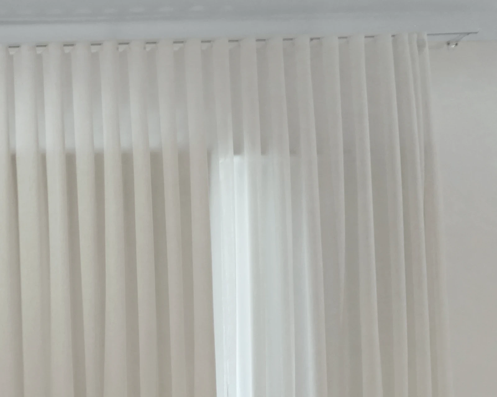 White sheer S-fold curtain hanging in even waves from a white ceiling track, with window light behind