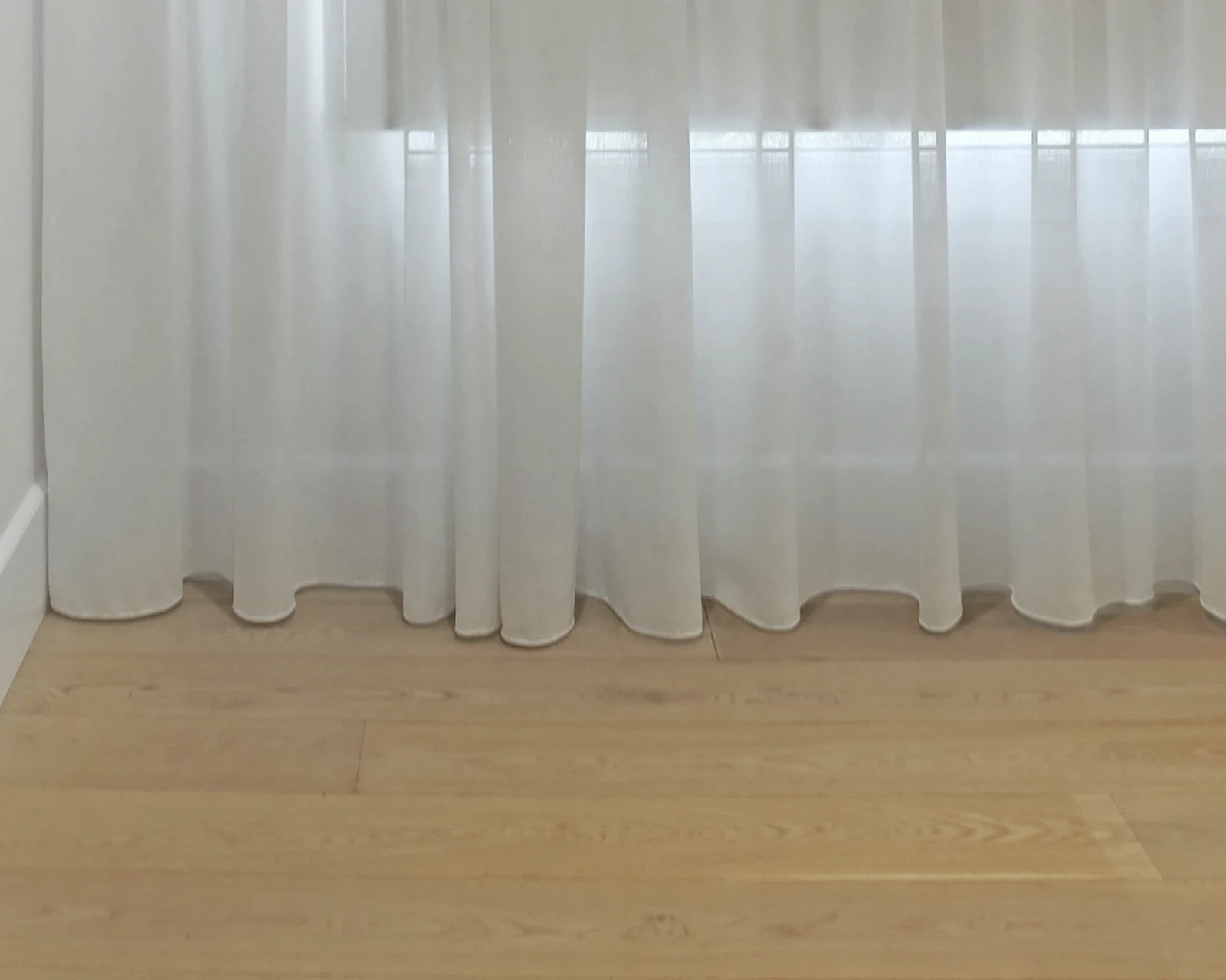 White sheer curtain hem just touching a timber floor, with daylight coming through the fabric