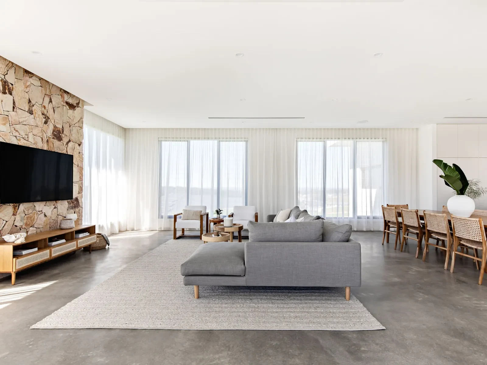 An open-plan living and dining room with ivory sheer curtains along a whole wall of windows, beside a stone feature wall An open-plan living and dining room with ivory sheer curtains along a whole wall of windows, beside a stone feature wall